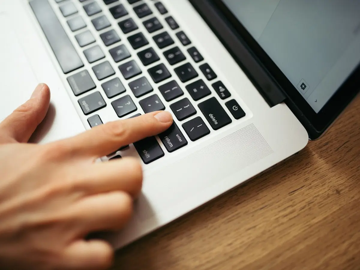 A hand pressing keys on a laptop keyboard resting on a wooden desk