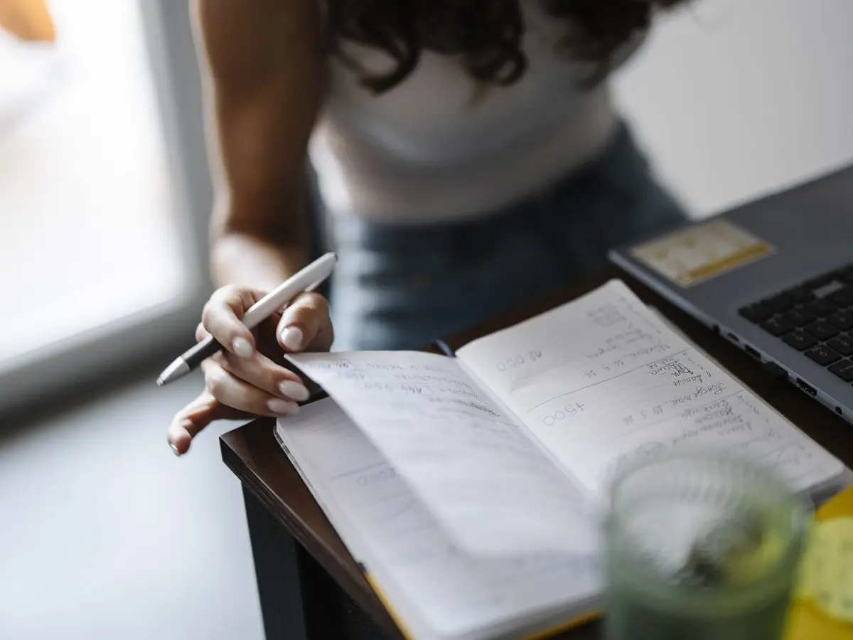 A person writing in an open notebook at a desk, with a laptop at the edge of the frame