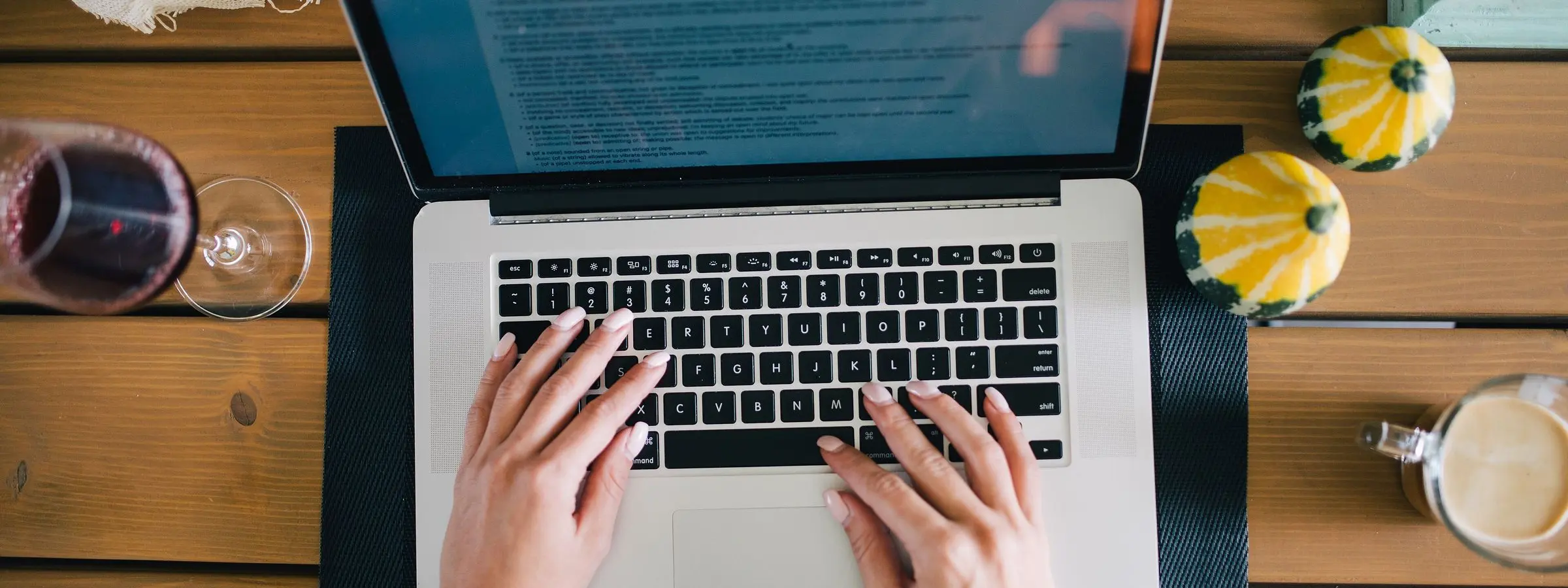 Hands typing on a laptop on a wooden table, seen from above, with a cup and small gourds beside it