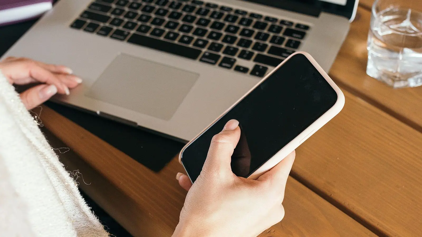 A hand holding a smartphone above an open laptop on a desk
