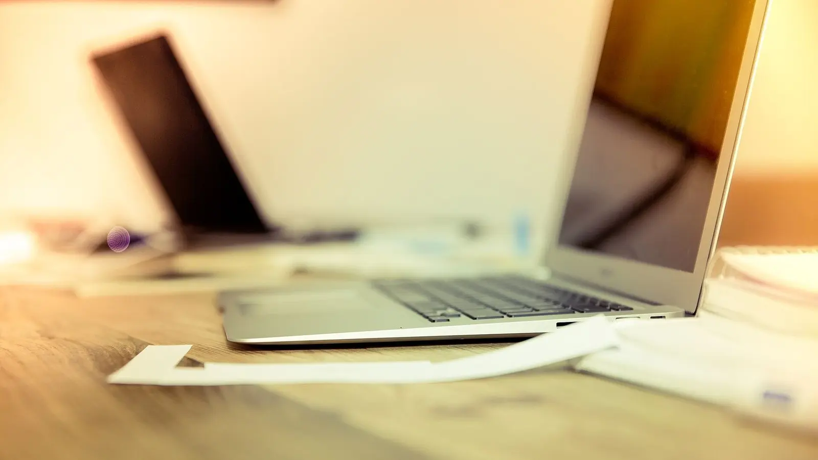 An open laptop on a wooden desk in soft golden light, with a sheet of paper in front of it