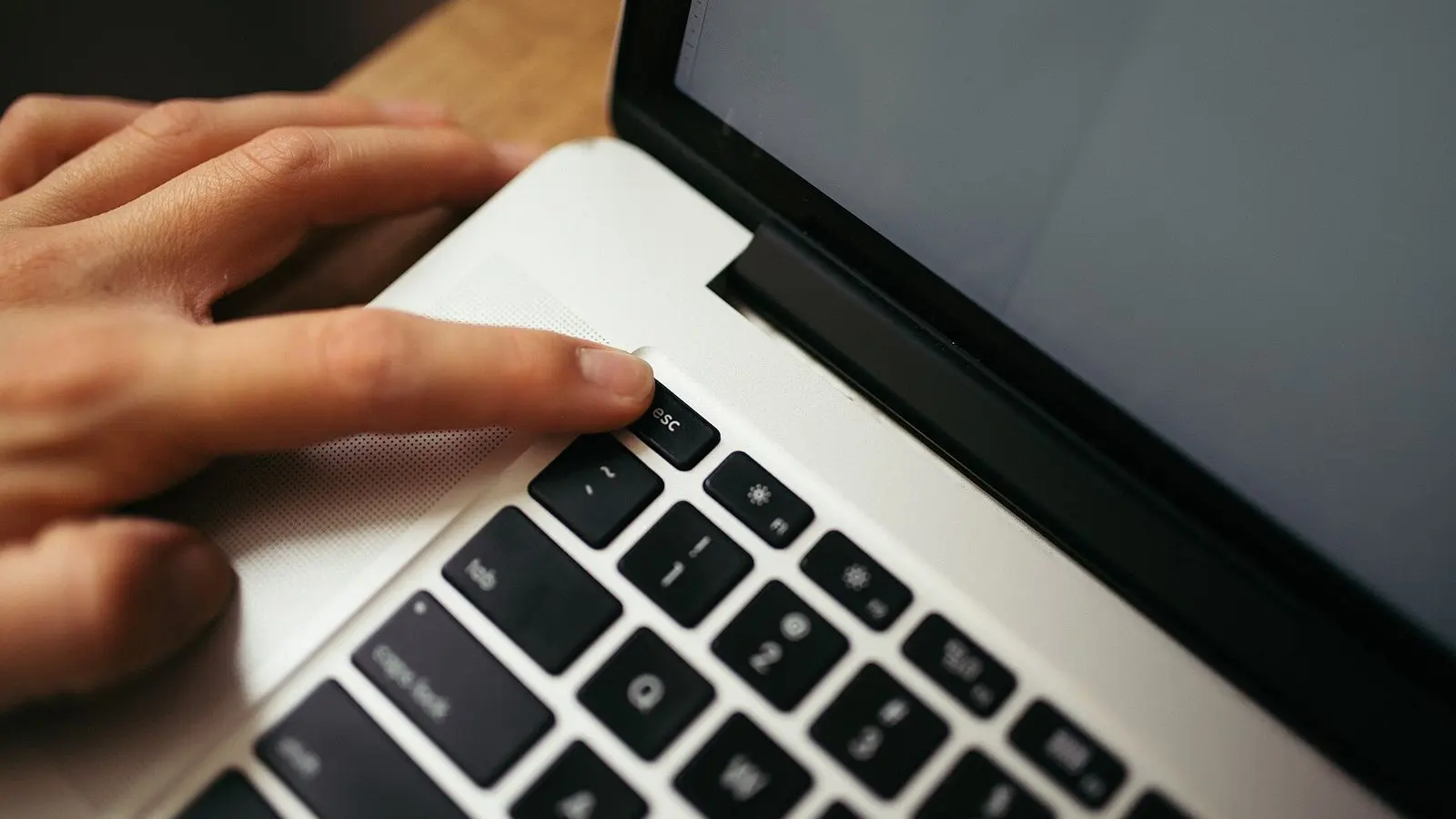 A hand resting on a laptop keyboard on a wooden desk, a finger pressing a key