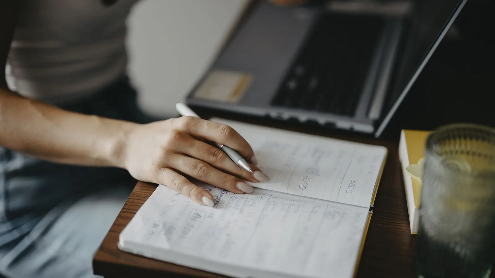 A hand writing notes in an open notebook beside a laptop on a desk, in soft daylight
