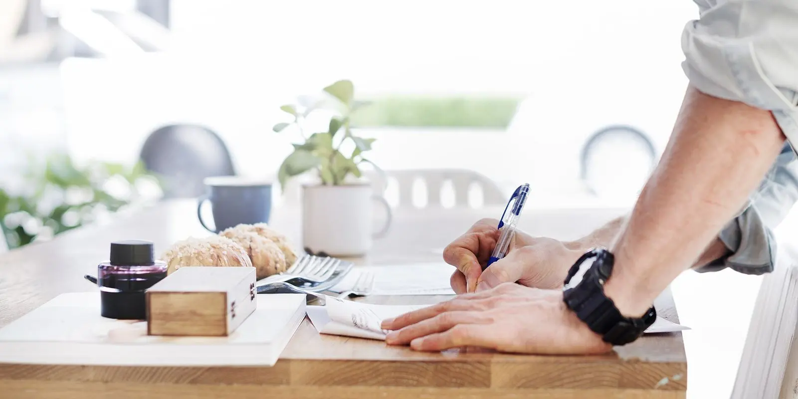 A hand writing on a sheet of paper at a white table, a cup and a small plant beyond