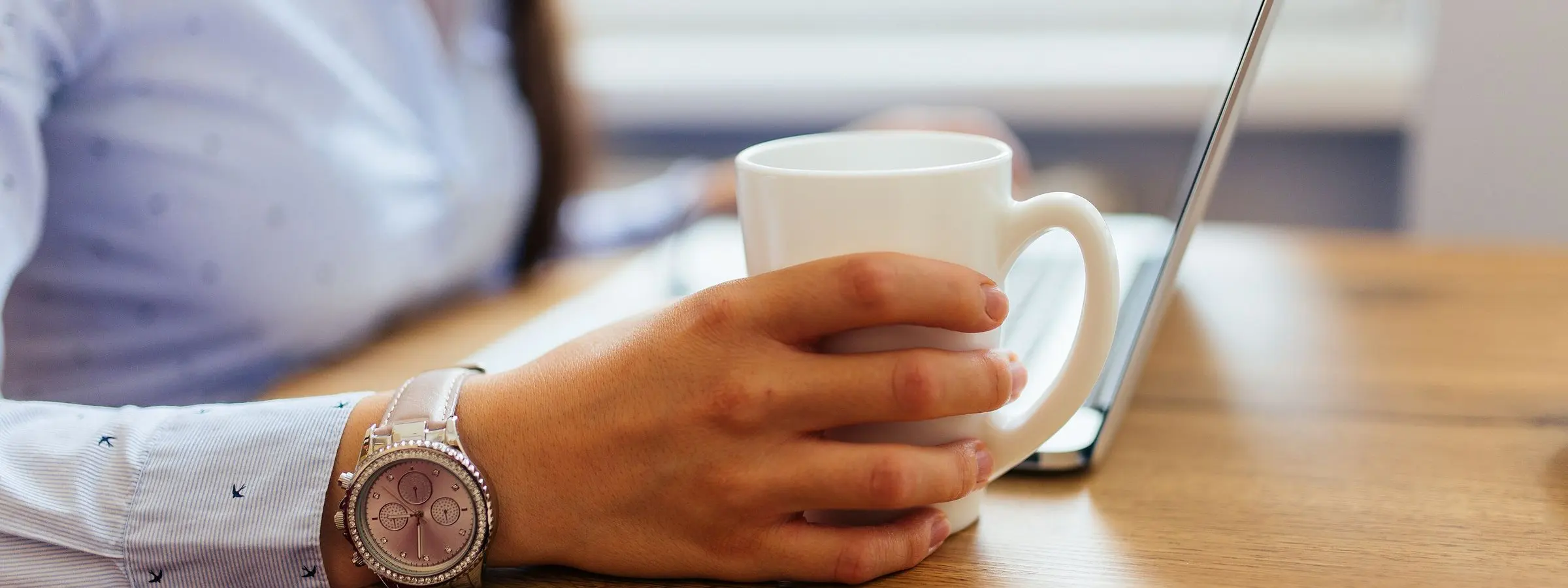A hand holding a white mug beside an open laptop on a warm wooden table