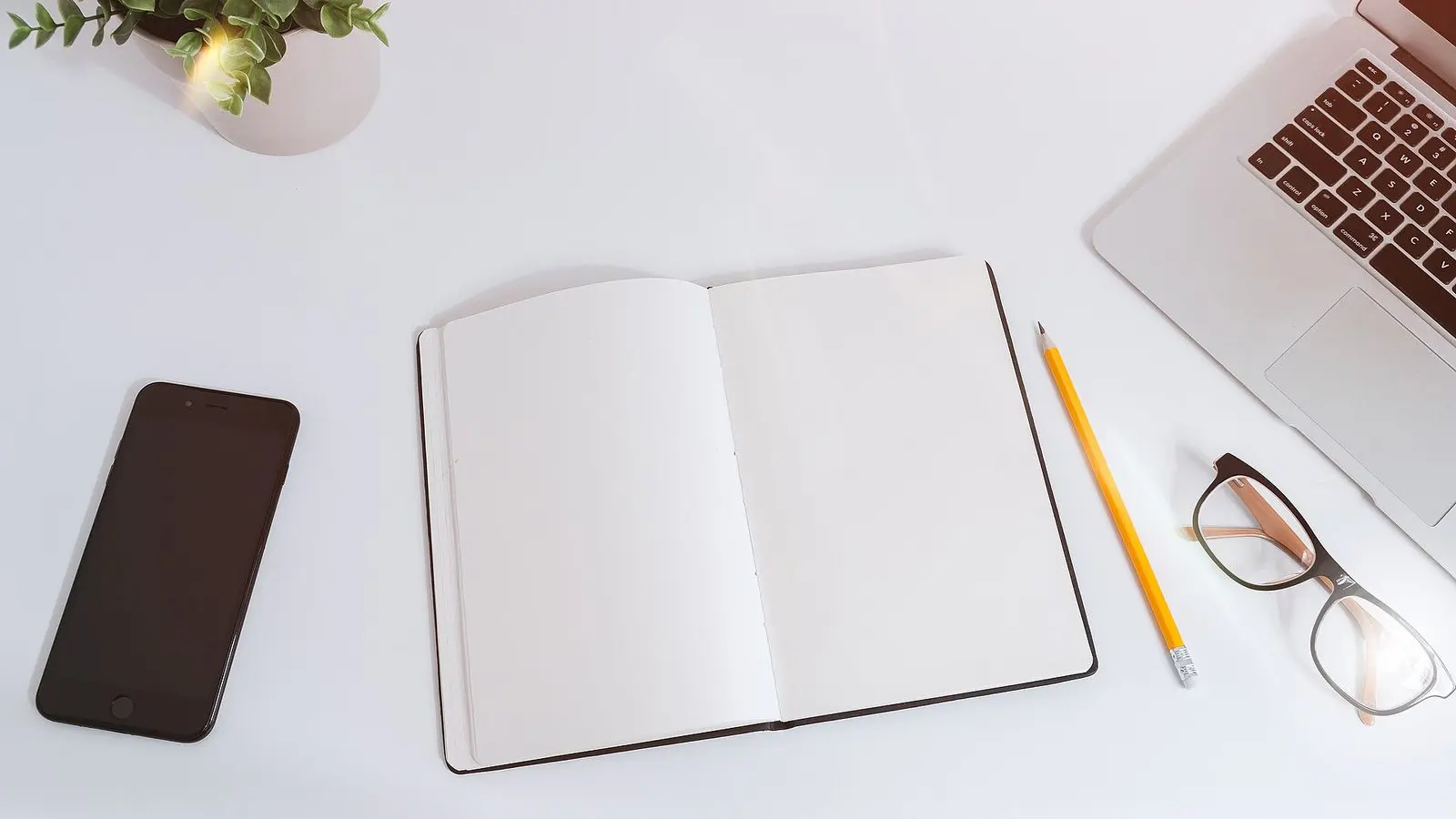 Seen from above, a smartphone, an open notebook, a pencil and glasses on a white table beside a laptop