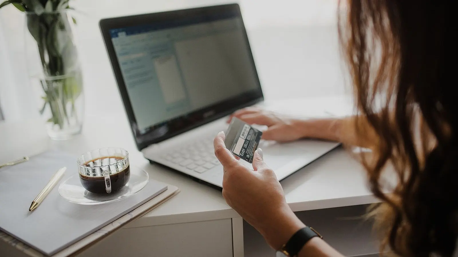 A person at a desk holding a payment card while typing on a laptop, a cup of coffee beside it