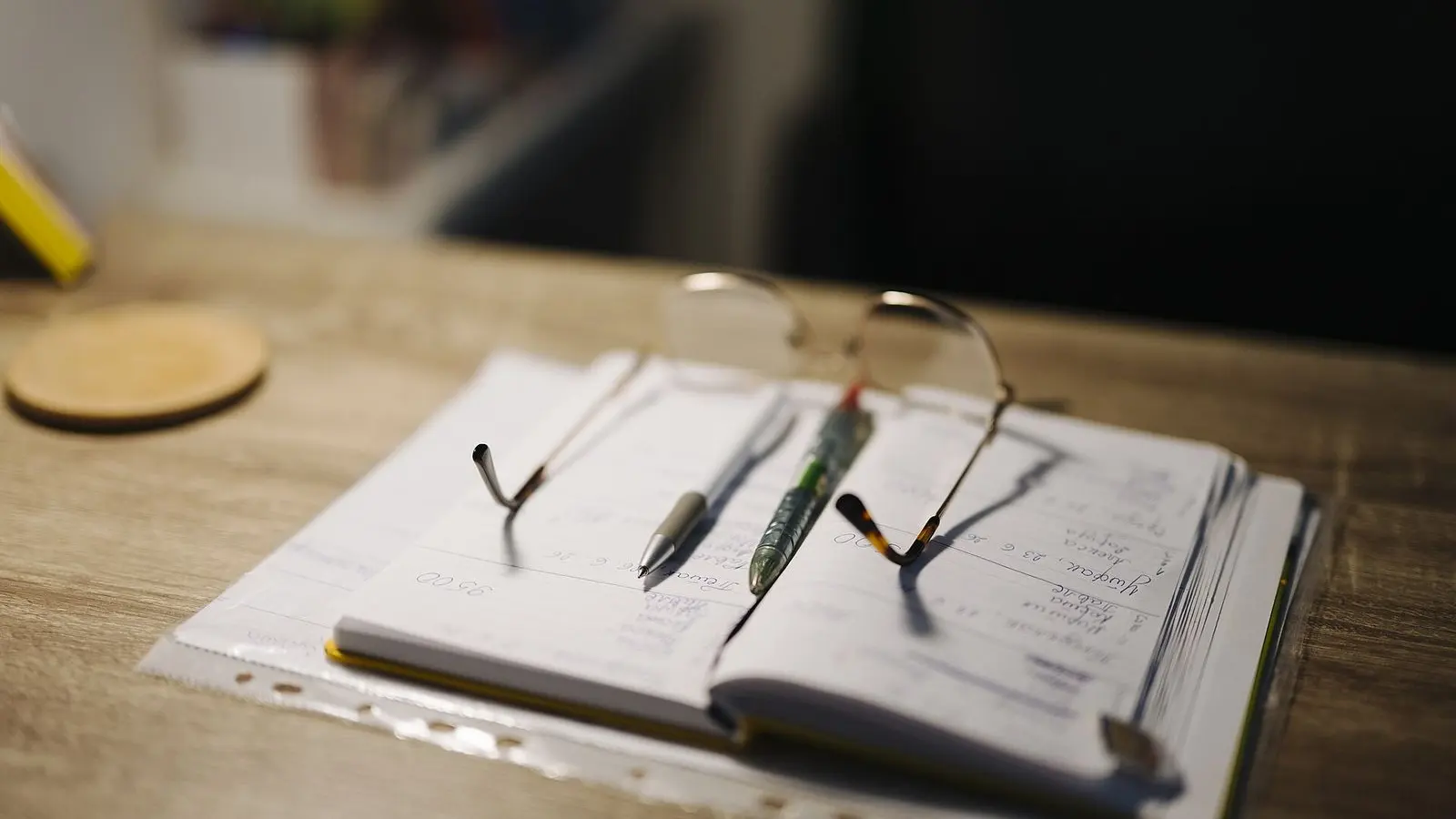 An open notebook with pens and a pair of reading glasses on a wooden desk