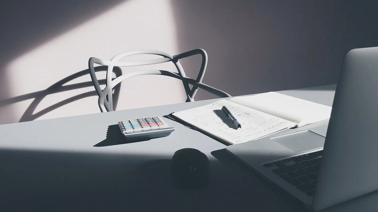 A calculator, a pair of glasses and a laptop on a desk, lit by low side light
