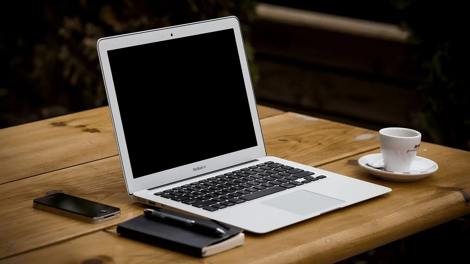 An open laptop on a wooden table beside a cup and saucer and a phone