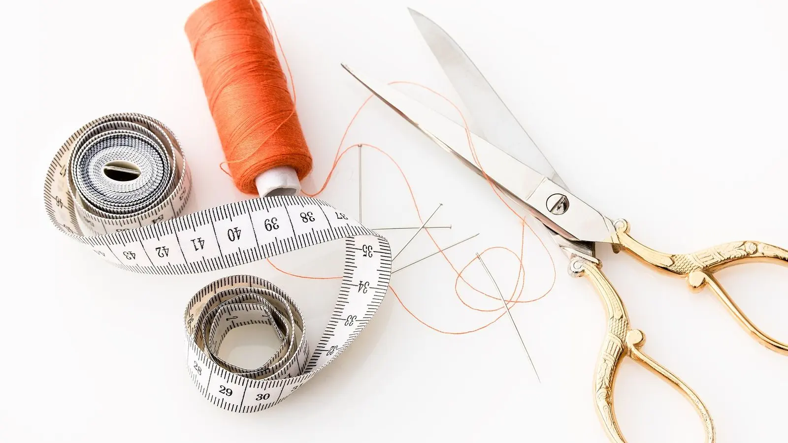 A tape measure, a spool of orange thread and a pair of scissors on a white surface