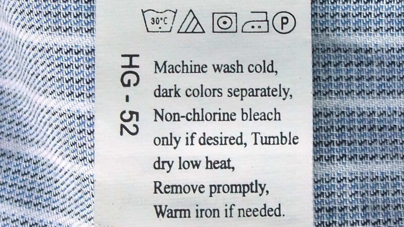A white care label with laundry symbols and washing instructions in English, sewn into a blue striped shirt