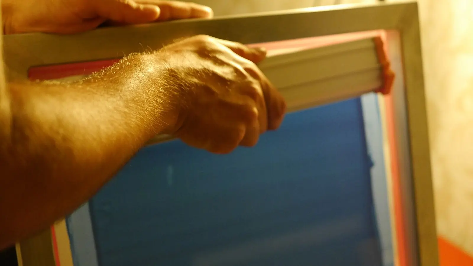 A hand drawing a squeegee across a screen-printing frame, ink spread on the mesh, in warm light