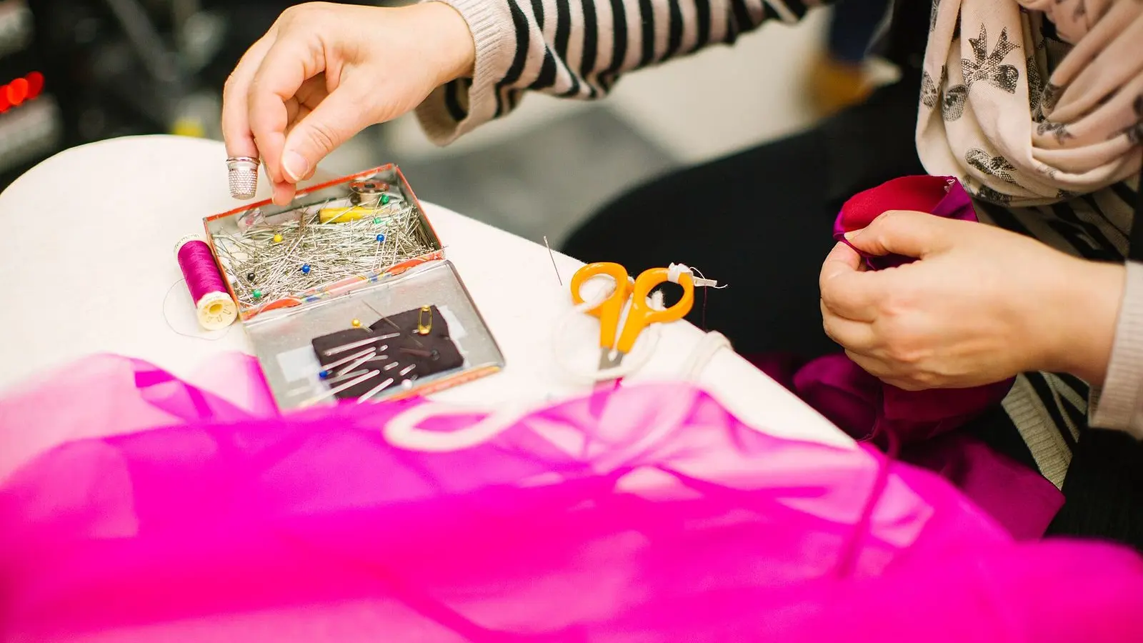 Hands working on a piece of bright pink fabric at a table, beside a tag and other sewing materials
