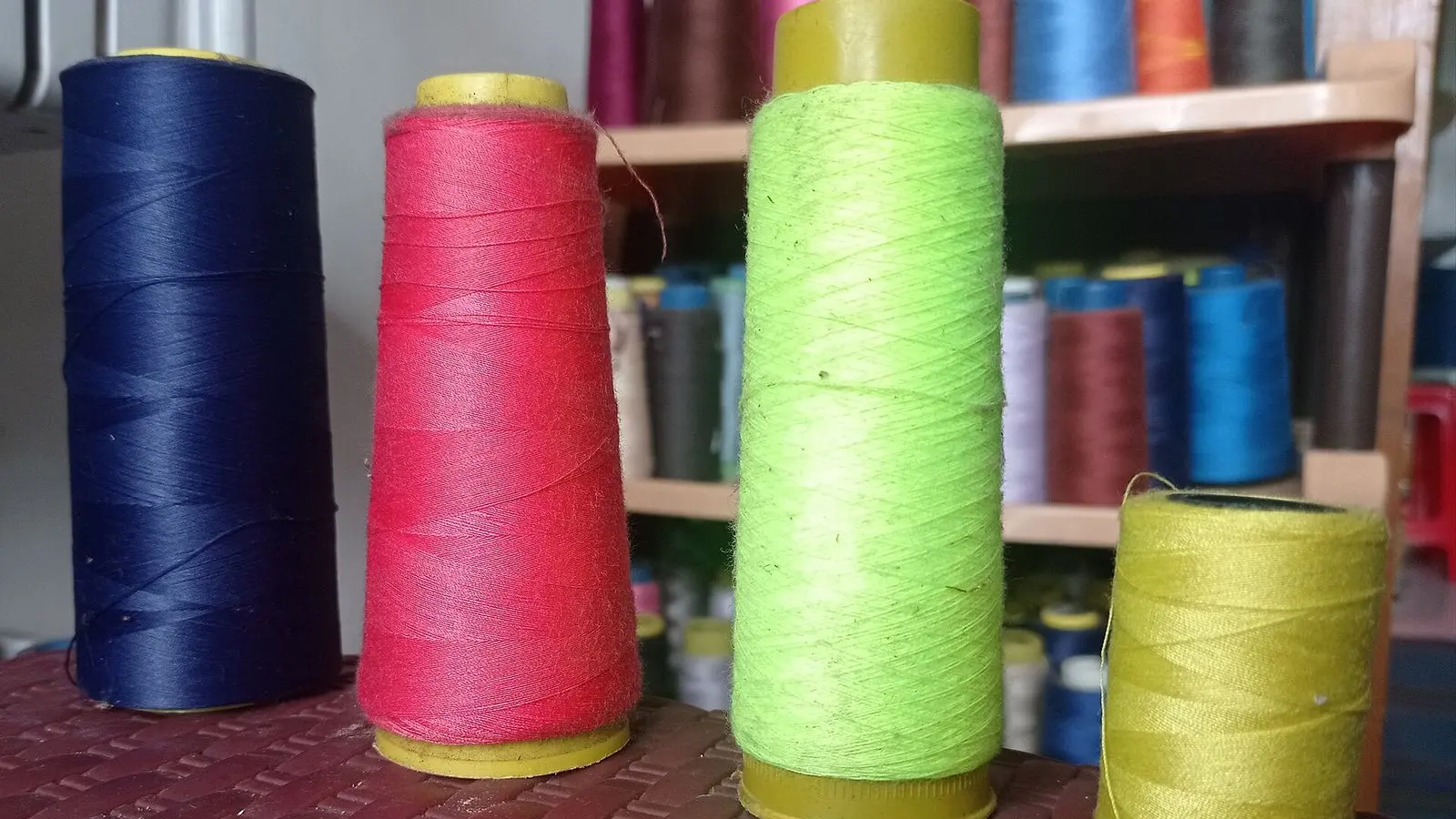 Cones of navy, red, green and olive sewing thread on a stool, with shelves of thread cones behind