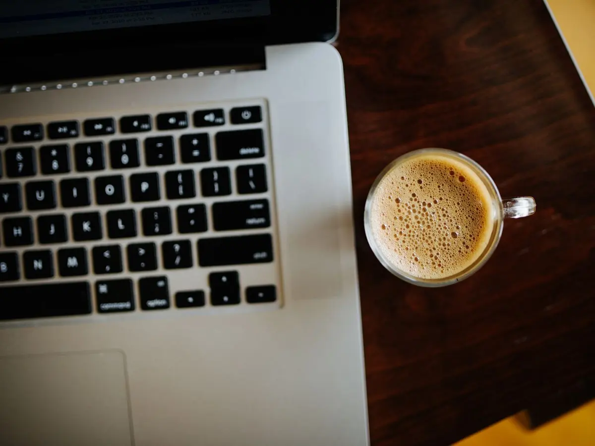 A cup of coffee beside an open laptop on a dark wooden table, seen from above