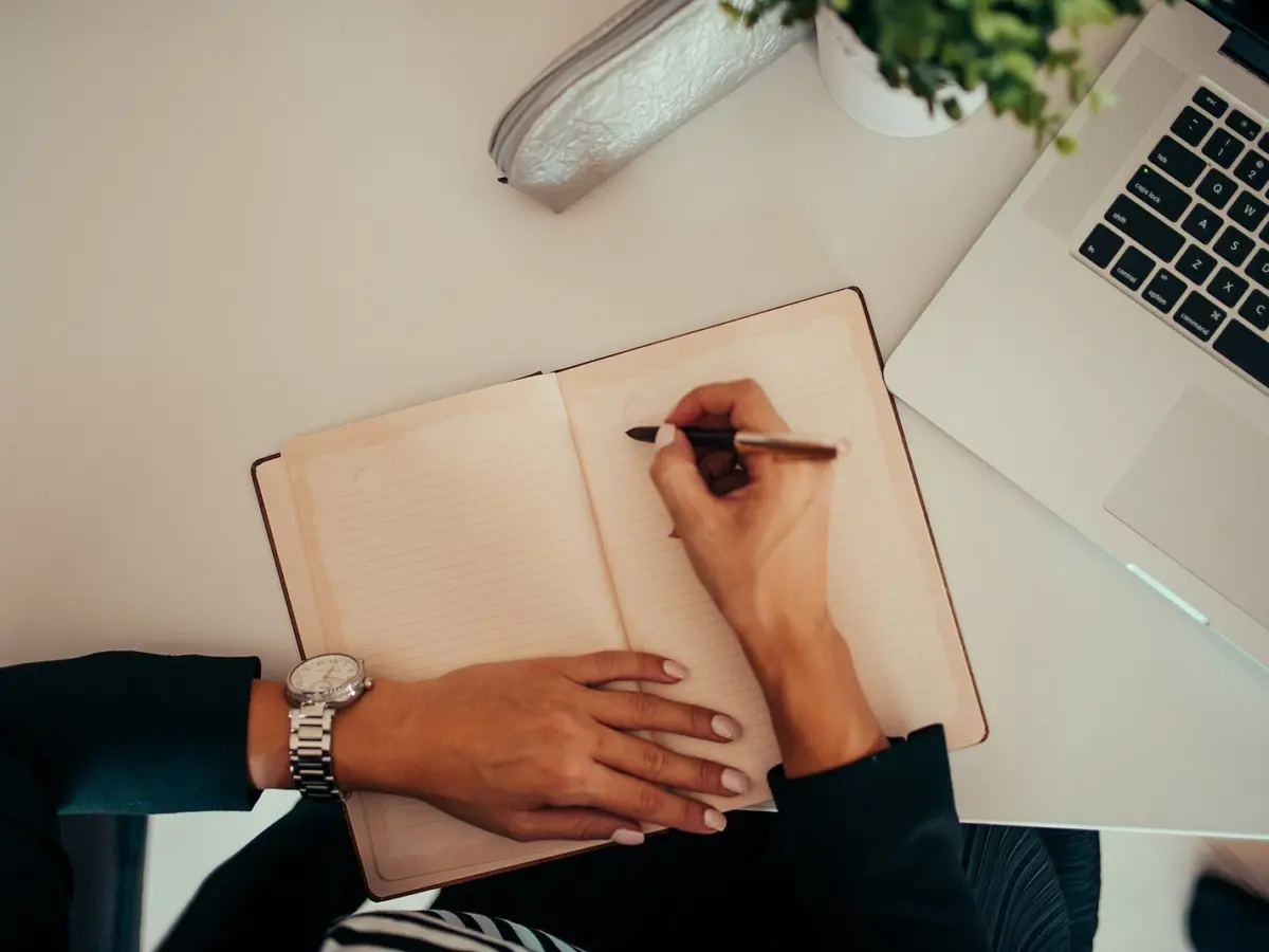 Hands writing in a notebook with a pen while a laptop sits open on the desk beside it