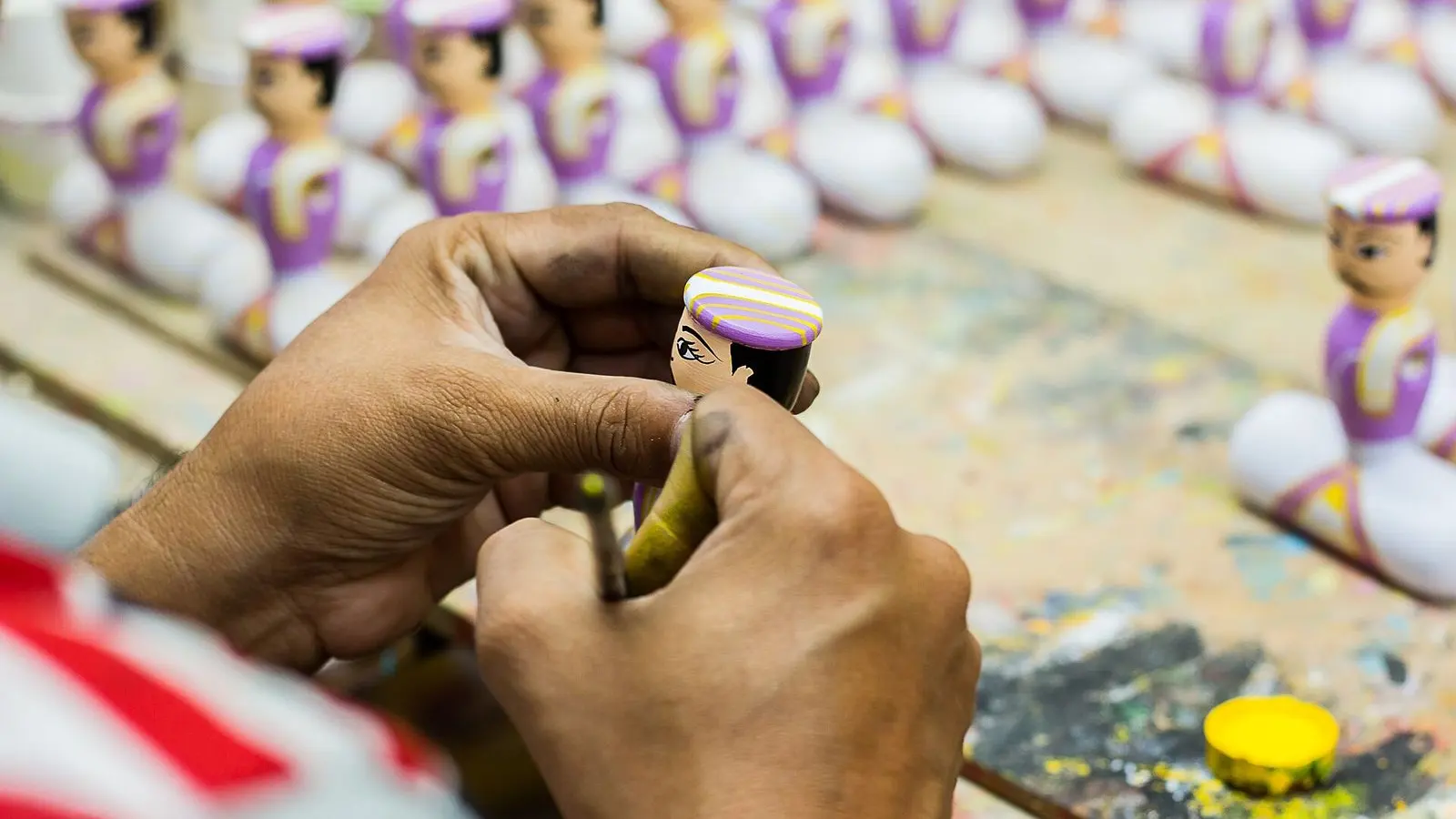Hands painting a small wooden Channapatna doll, with rows of finished dolls on the workbench behind