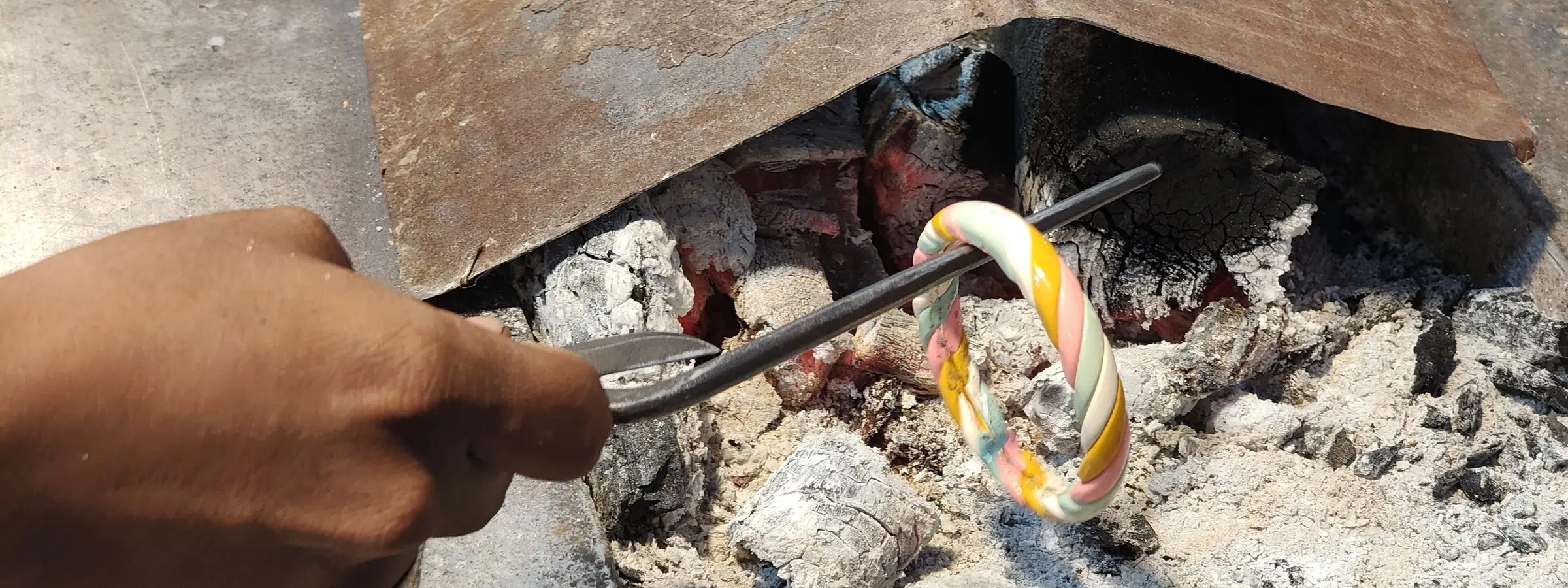 A hand holding a lac bangle over hot coals with tongs at a bangle maker's bench in Laad Bazaar, Hyderabad