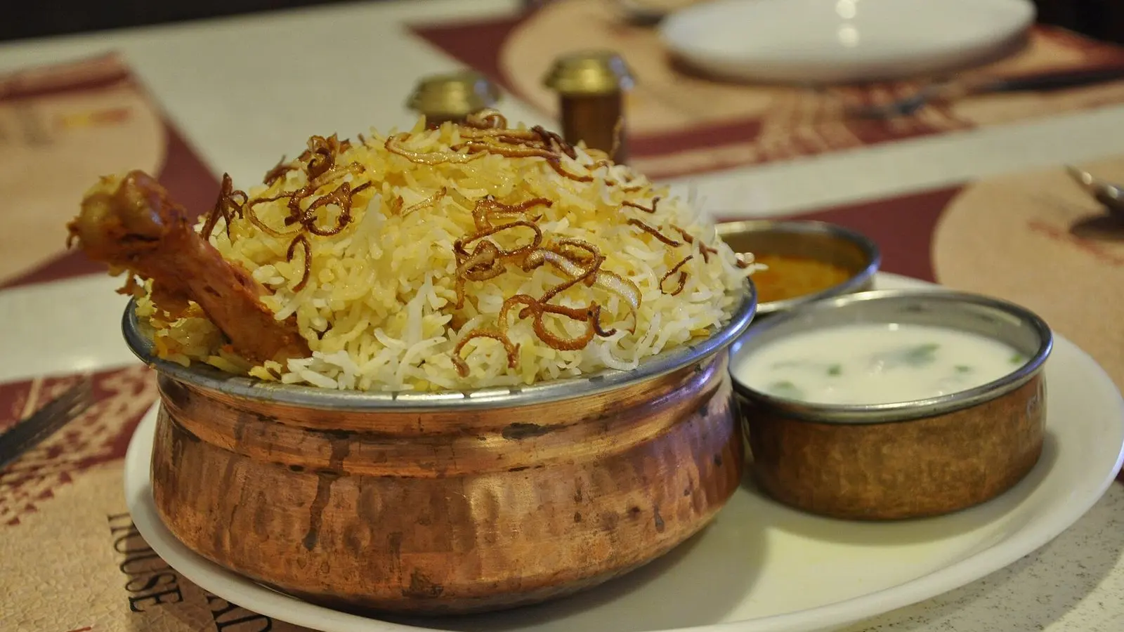 Hyderabadi chicken biryani served in a copper handi beside a bowl of raita on a table