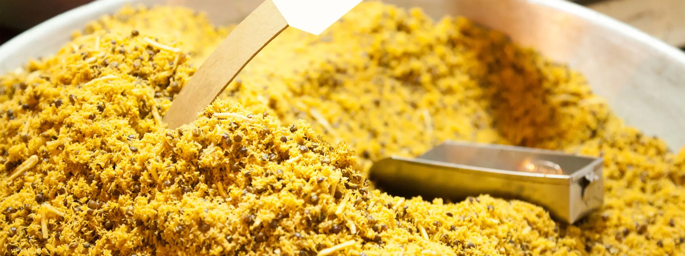Yellow namkeen heaped in a steel tray with a metal scoop and a blank price tag