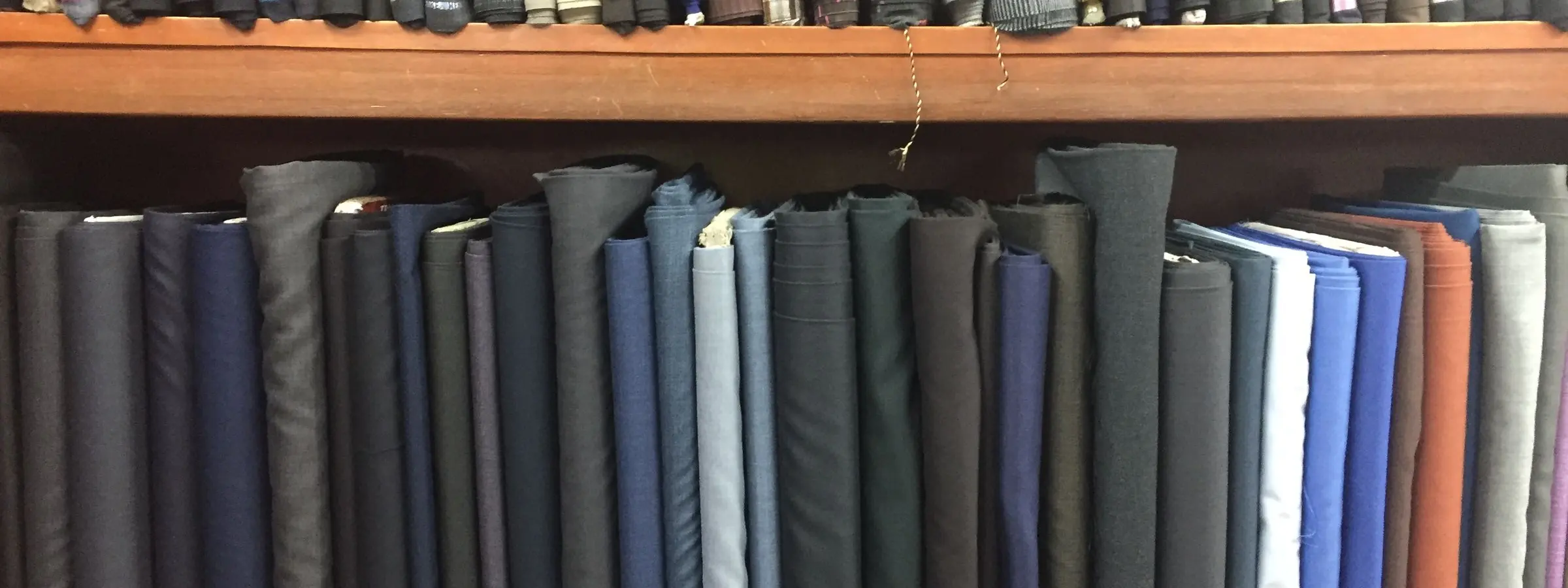 Bolts of suiting fabric in shades of grey, blue and brown stacked on shelves in a Mumbai tailor's shop