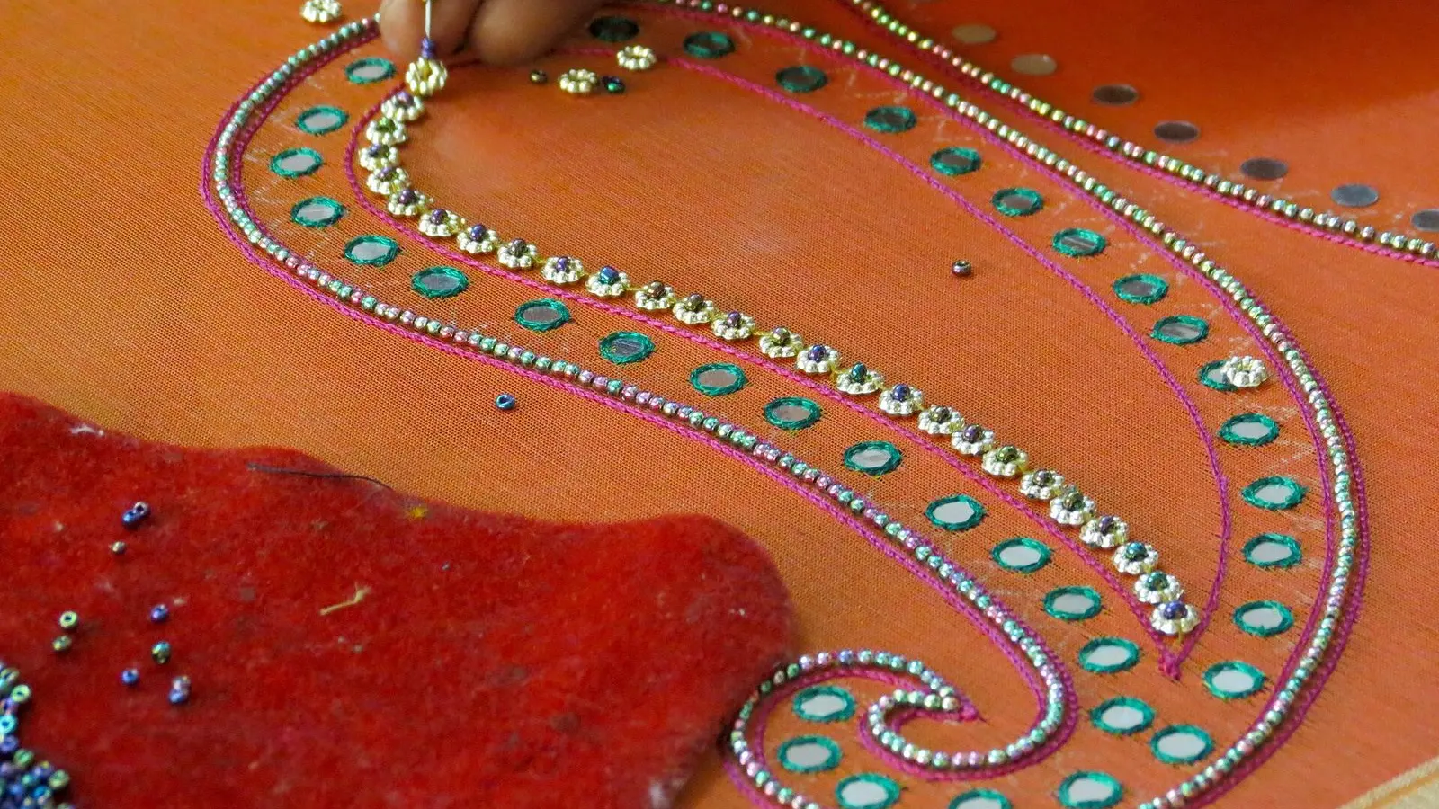 Close view of zari and mirror work being stitched onto orange fabric by hand