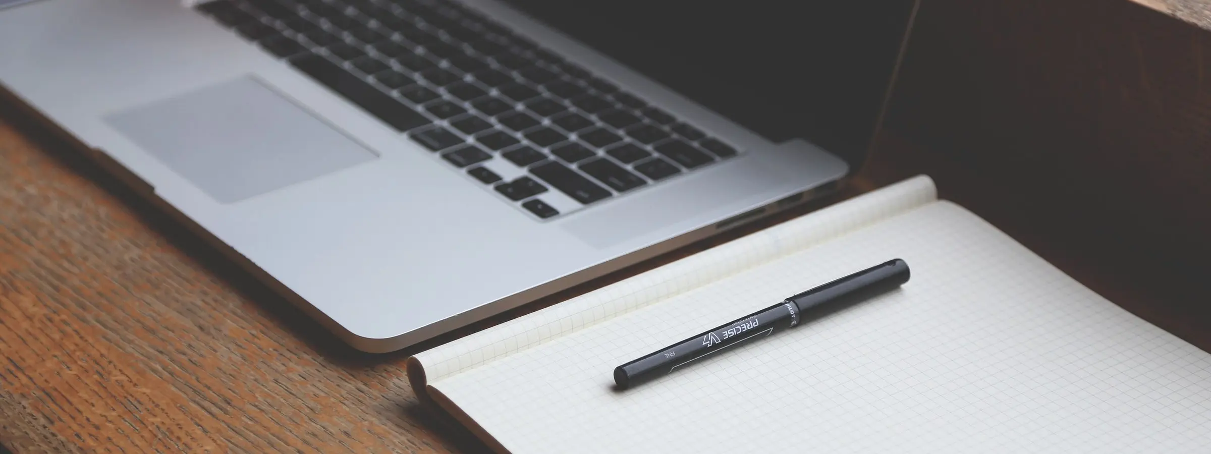 A silver laptop and an open notebook with a pen on a wooden table