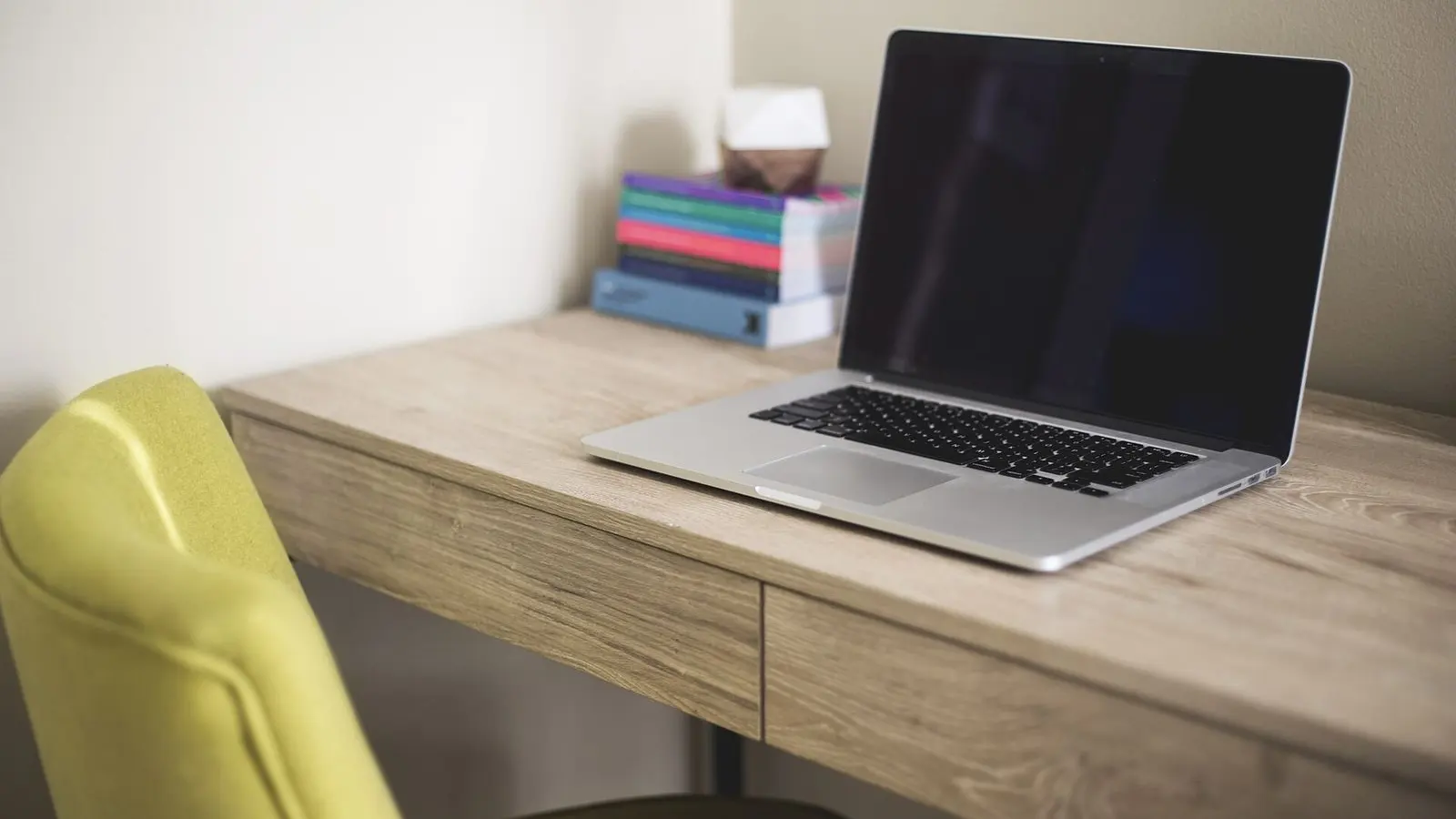 An open laptop on a light wooden desk beside a pile of books, with a chair pulled up