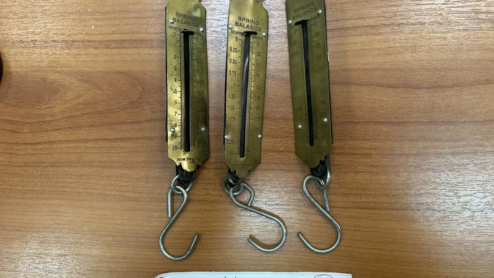Three brass spring balances with hooks lying side by side on a wooden surface, a handwritten tag beside them