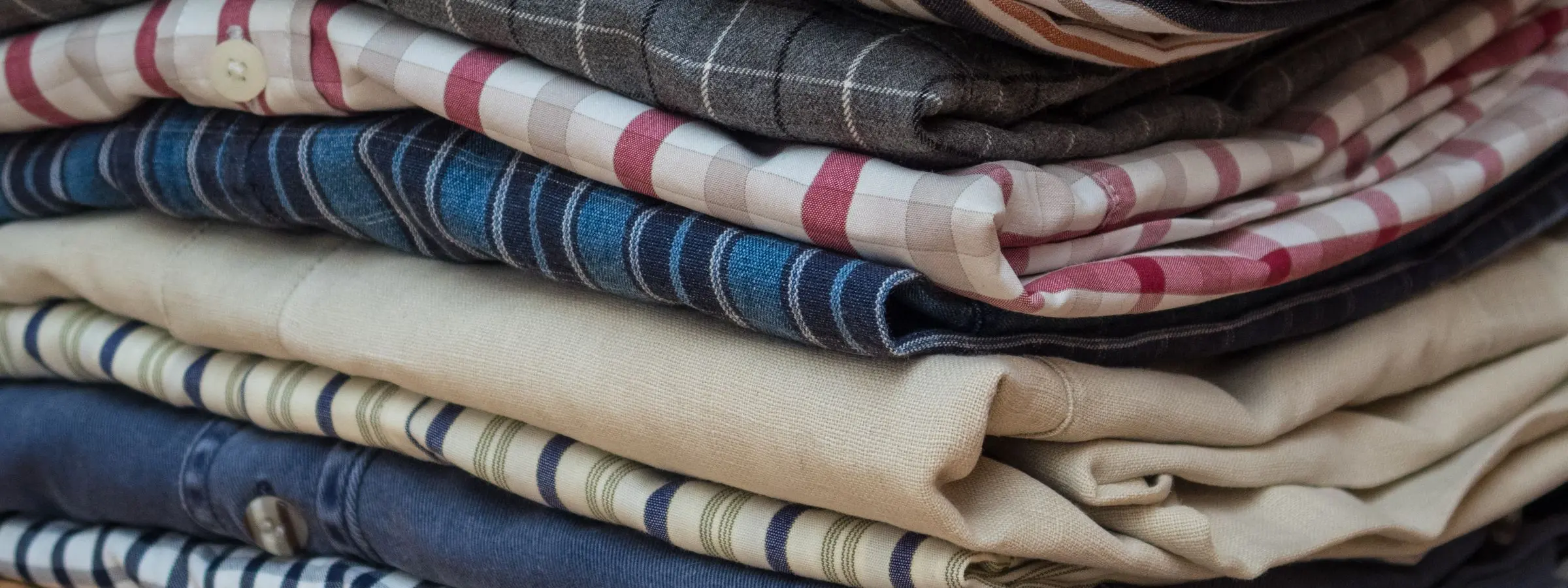 A tall stack of folded, ironed men's shirts in striped and checked cotton on a wooden surface