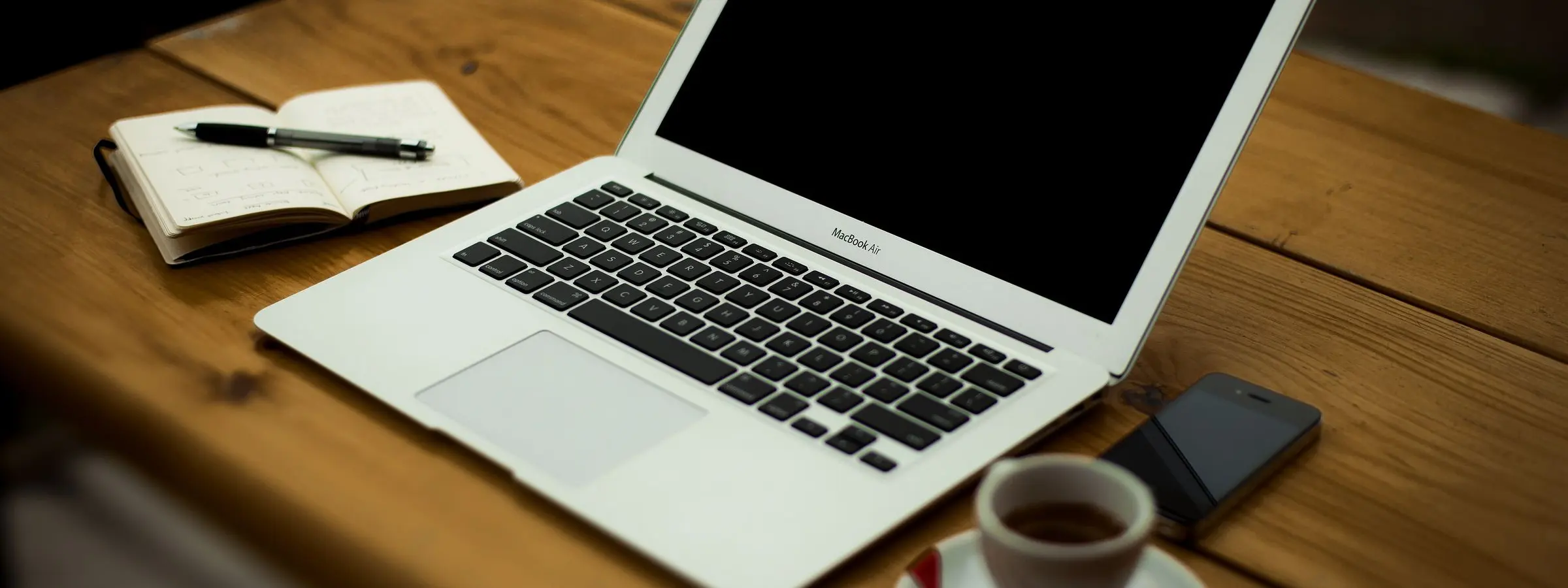 An open laptop on a wooden desk with a notebook, a pen, a phone and a cup of coffee