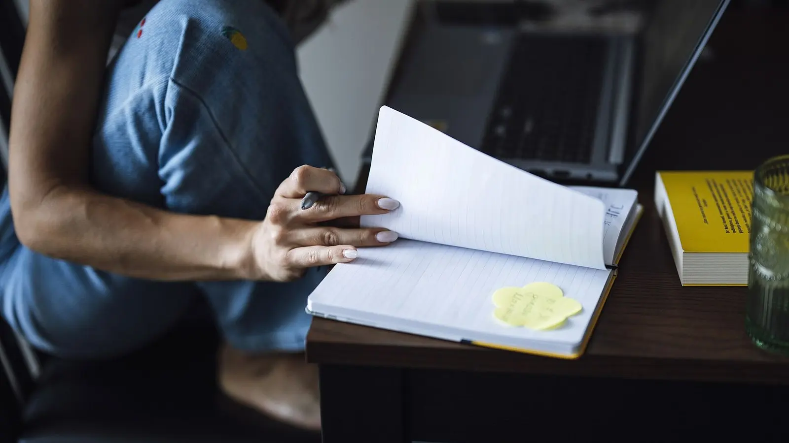 A person at a desk turning the pages of a notebook of handwritten notes, beside a laptop