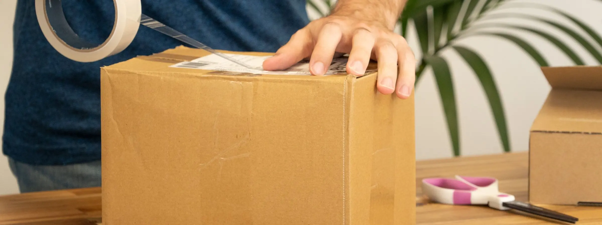 Hands taping a shipping label onto a cardboard parcel on a wooden table