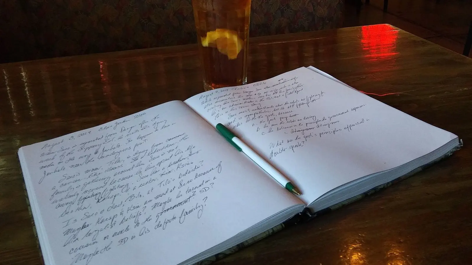An open notebook of handwritten notes with a pen, beside a glass of iced tea on a dark wooden table