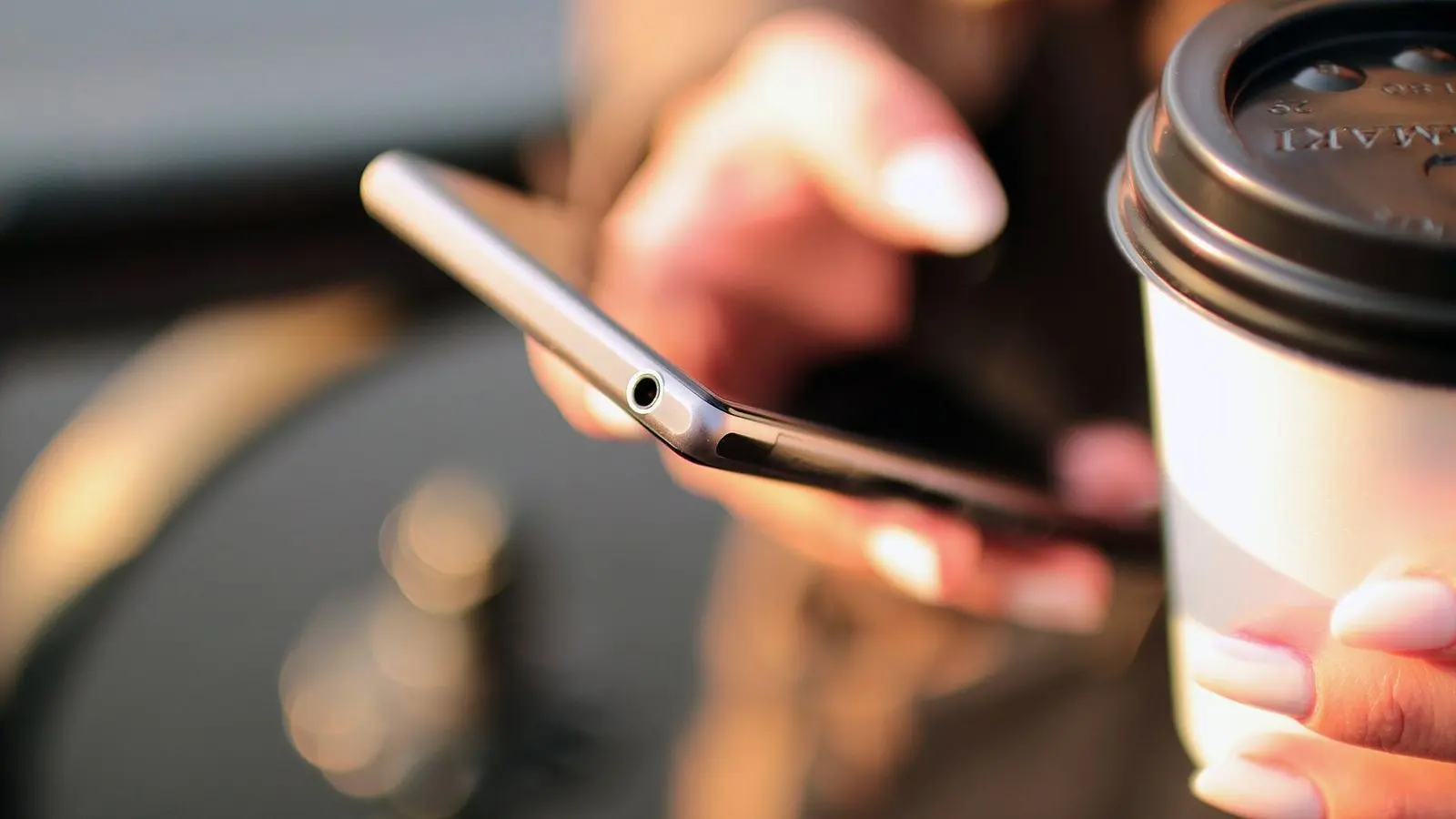 A hand holding a smartphone beside a paper coffee cup in soft warm light