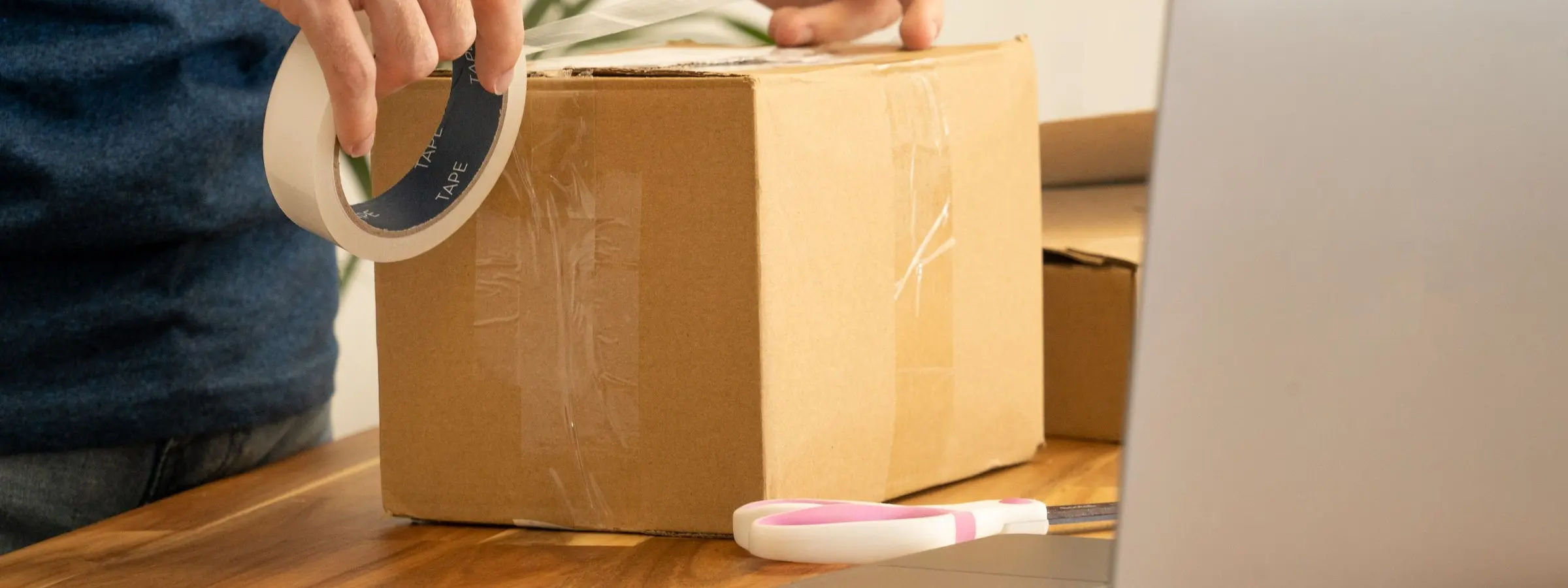 Hands sealing a cardboard parcel with tape beside an open laptop on a wooden table