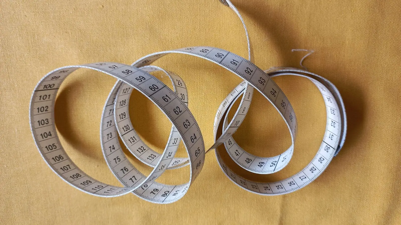 A white cloth tape measure in loose coils on mustard-yellow cotton