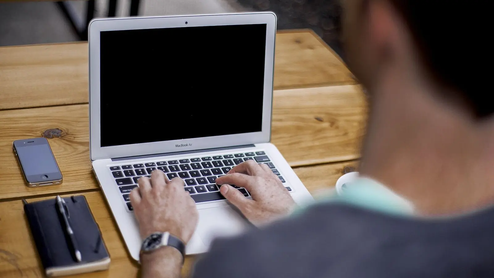 Hands typing on a laptop on a wooden desk, with a phone and a closed notebook beside it