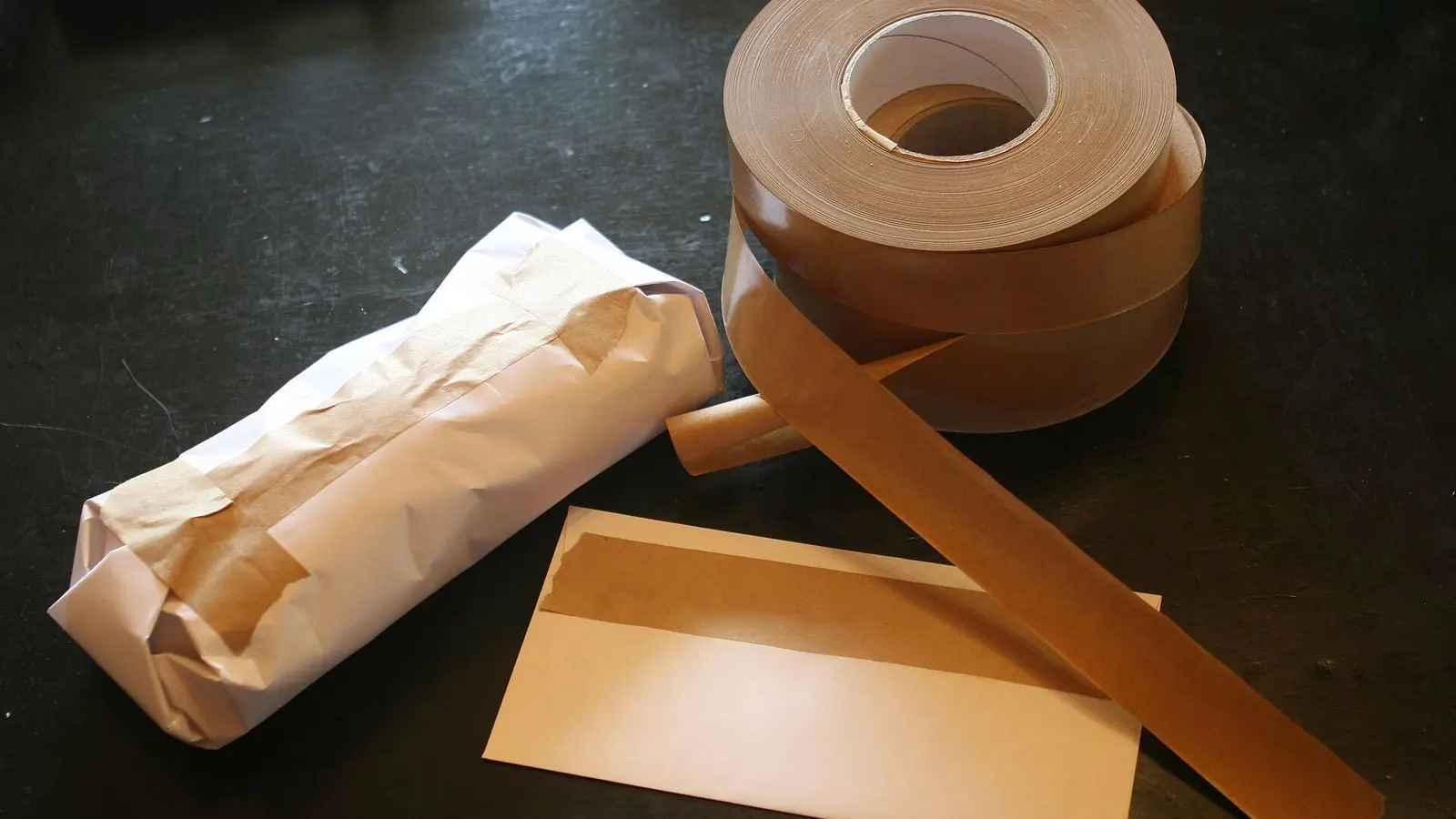 A roll of brown gummed paper tape beside a small parcel wrapped in kraft paper and a brown envelope