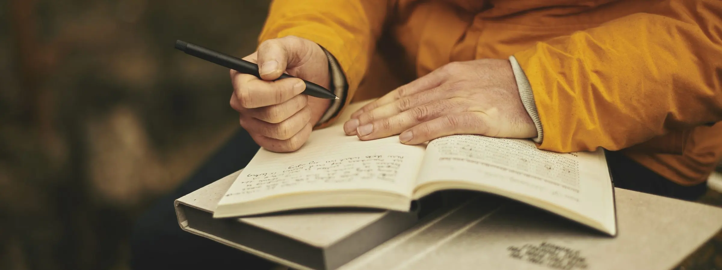 A person in a mustard jacket writing with a pen in an open notebook on their lap