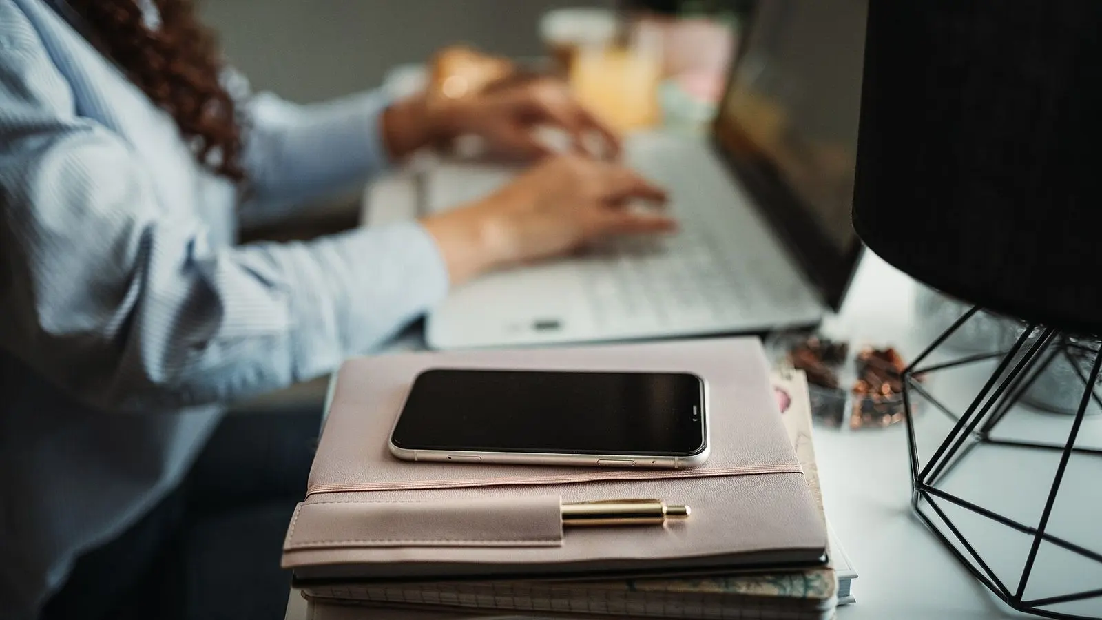 Hands typing on a laptop beside a smartphone resting on a pile of notebooks