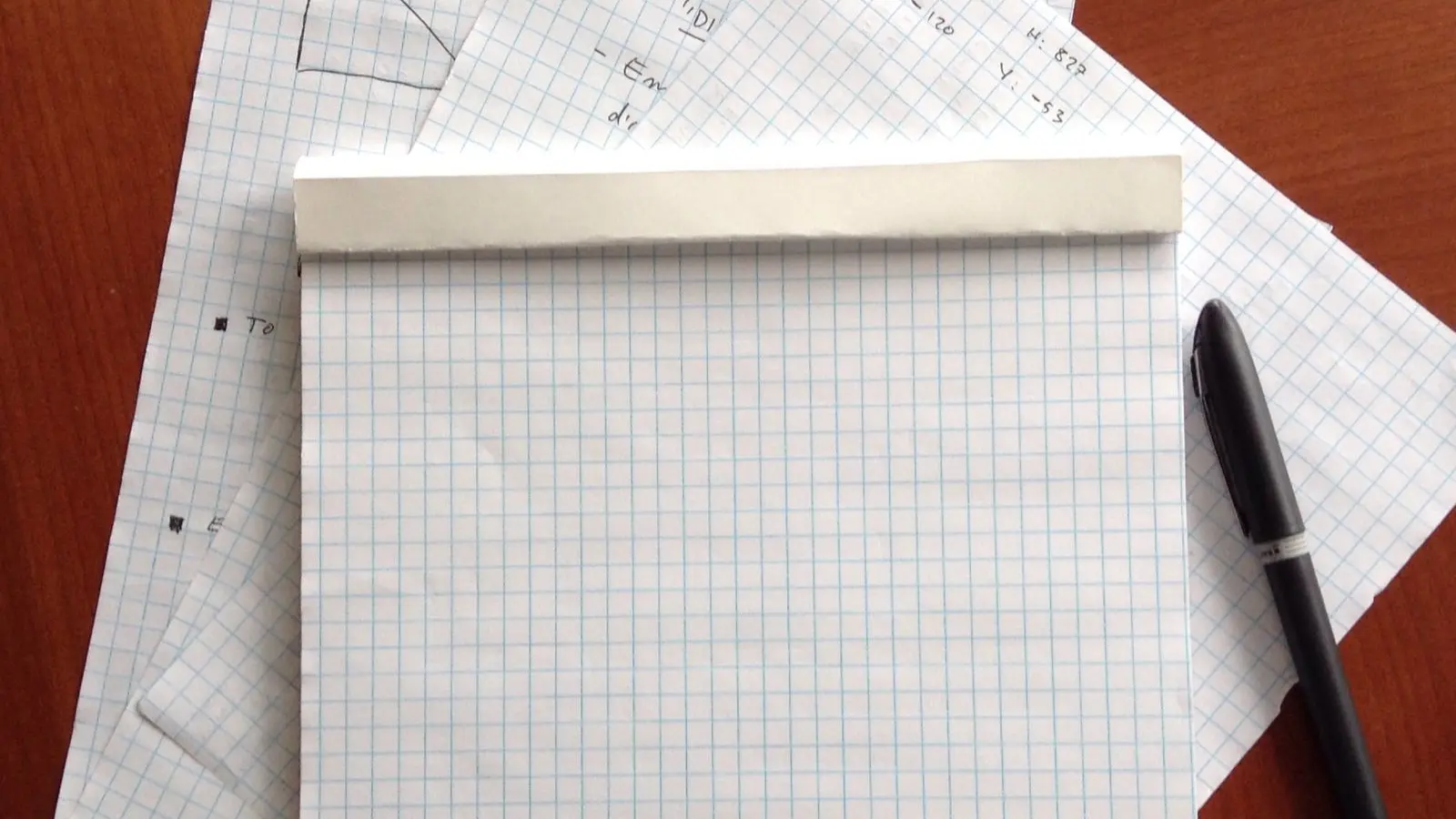 A pad of squared paper and a pen on a wooden desk, over sheets with a hand-drawn graph and sums