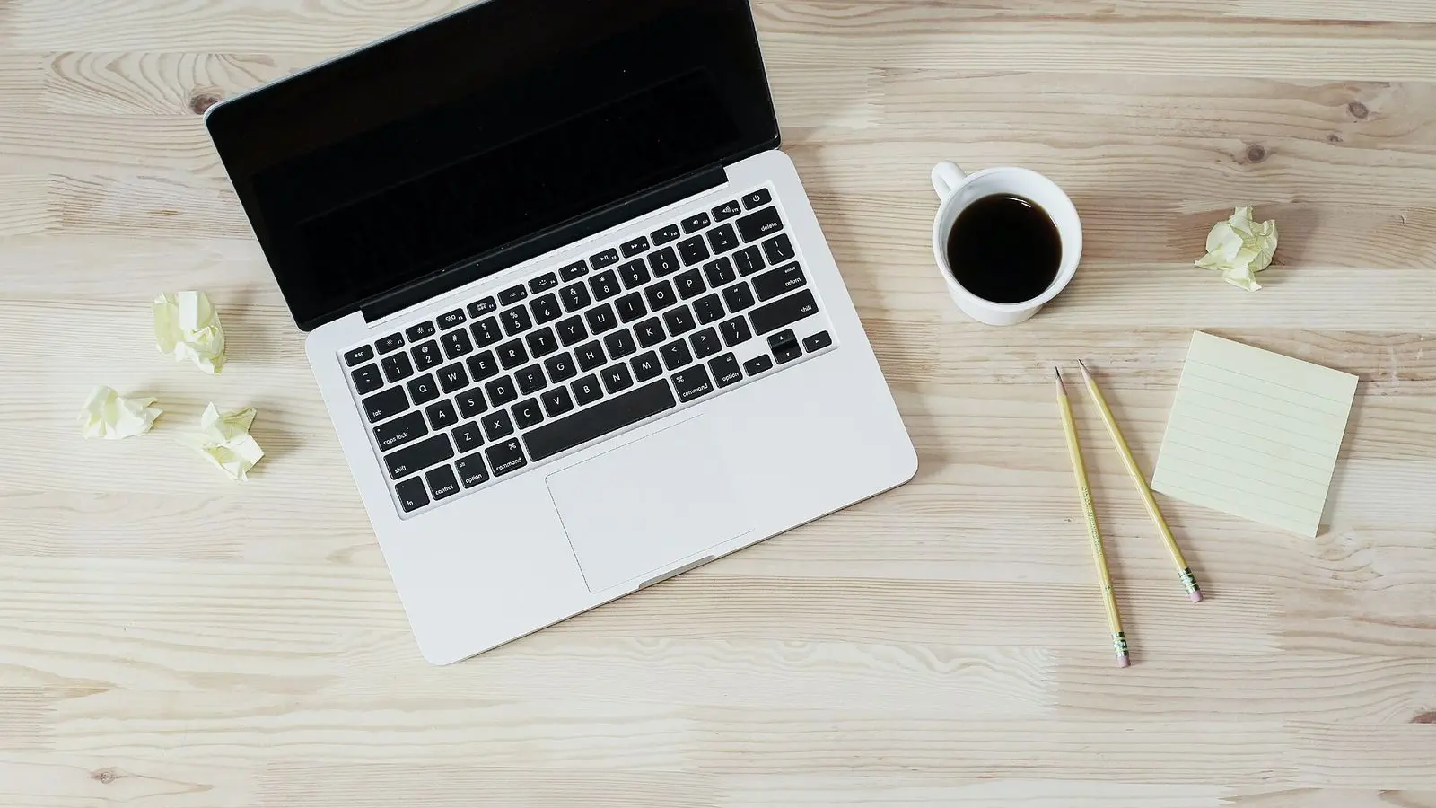 An open laptop, a cup of black coffee and two pencils on a pale wooden table