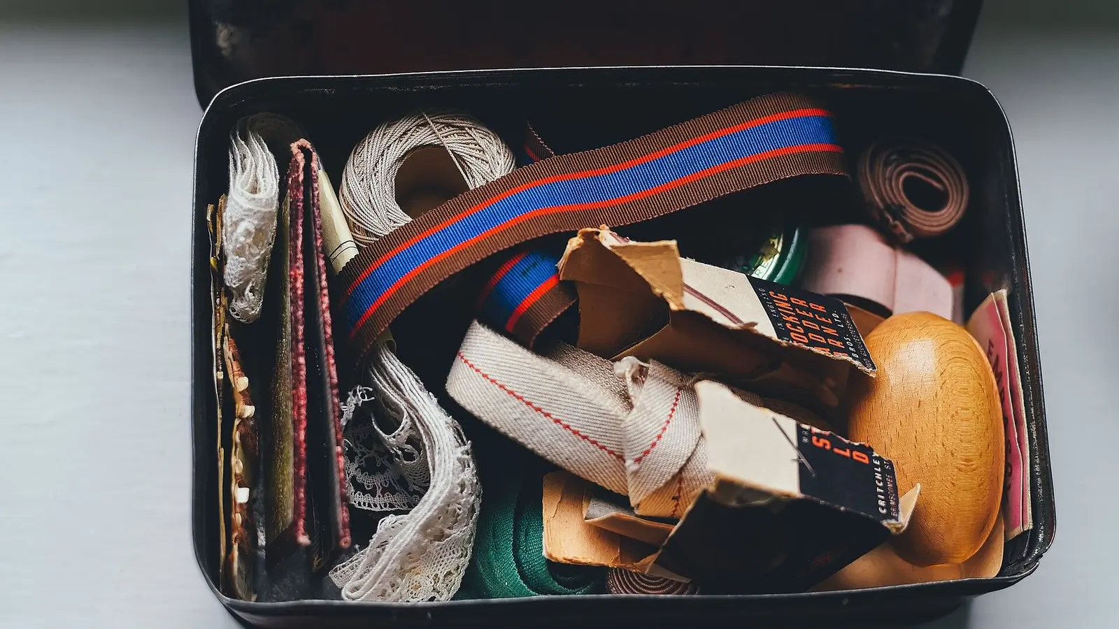 An open tin sewing box filled with ribbons, tape, bobbins and folded trims