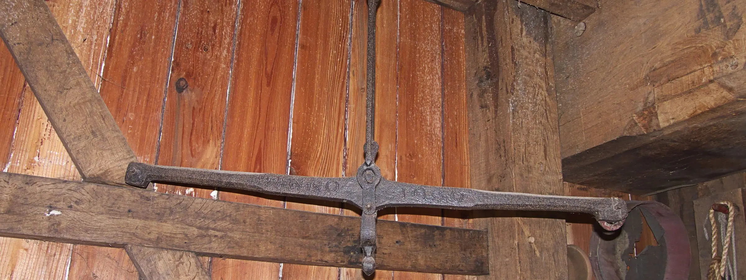 An iron balance beam hanging level from a hook against the wooden planks of a windmill