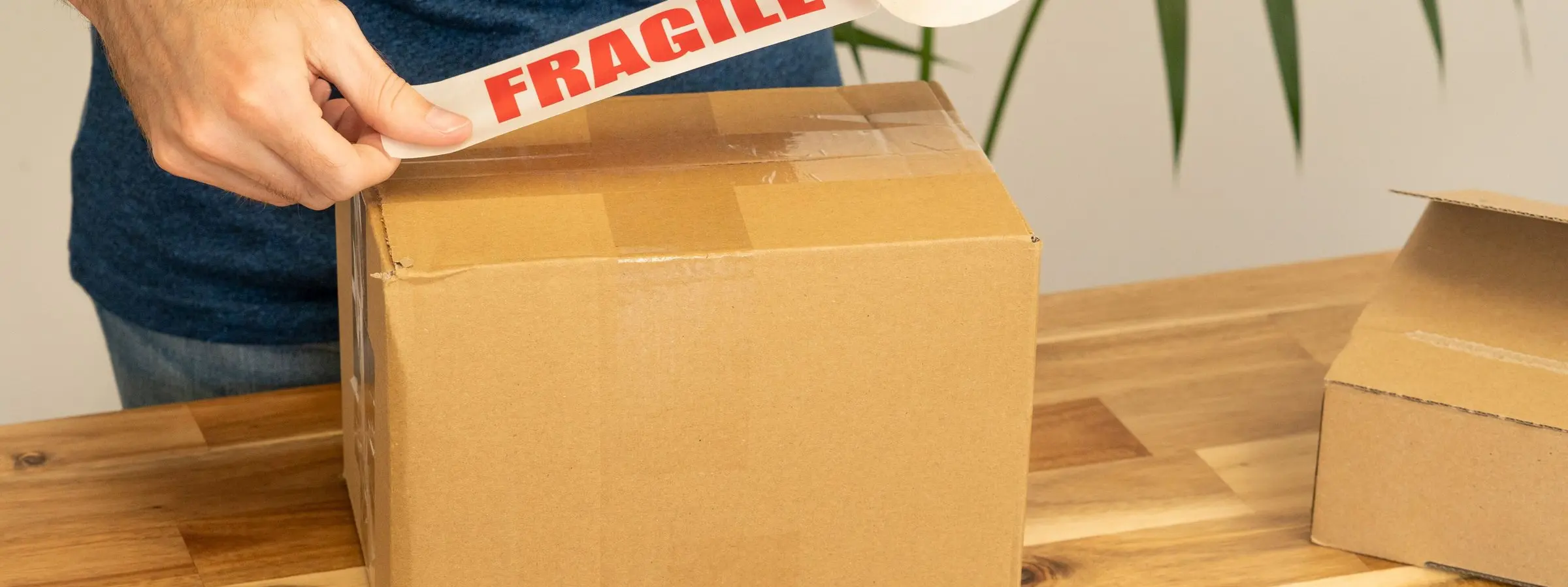 Hands sealing a cardboard box with red and white fragile tape on a wooden table